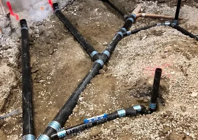 Network of black plumbing pipes laid in a gravel trench, connecting at various angles, with some marked in red and blue.
