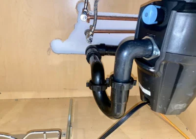 A close-up of plumbing under a sink, featuring a black pump connected to copper pipes and various hoses in a wooden cabinet.