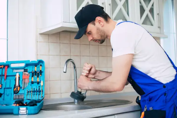 services 1 webp new A maintenance worker in a blue uniform uses a plunger at a kitchen sink, with a tool kit filled with various tools beside him.